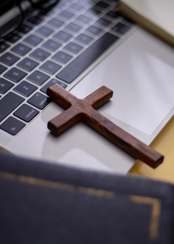 Wooden cross lying on a laptop keyboard with a closed book in the foreground.