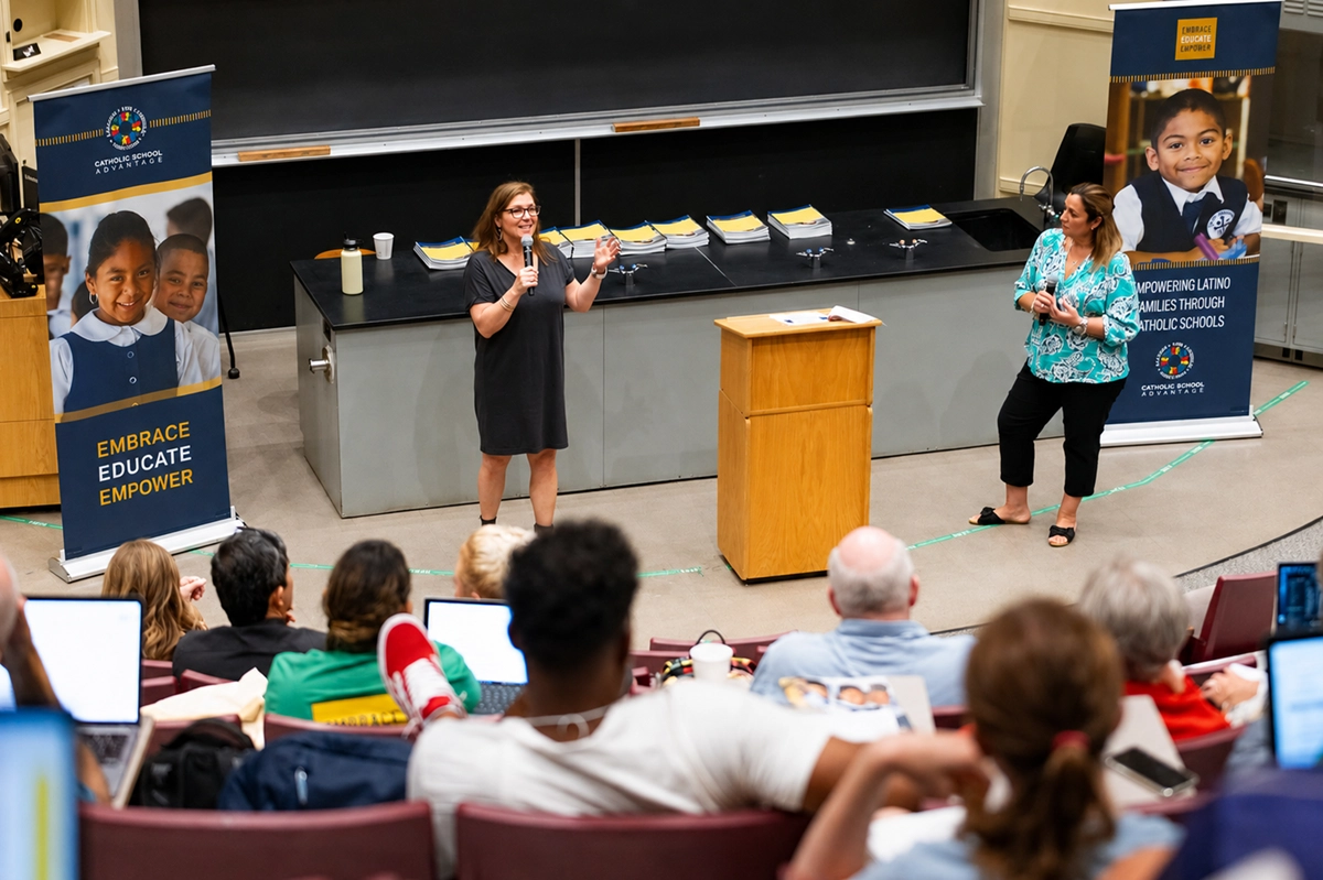 Kristin Moore presenting to a full lecture hall of Catholic school leaders, microphone in hand, beside Embrace Educate Empower banners.