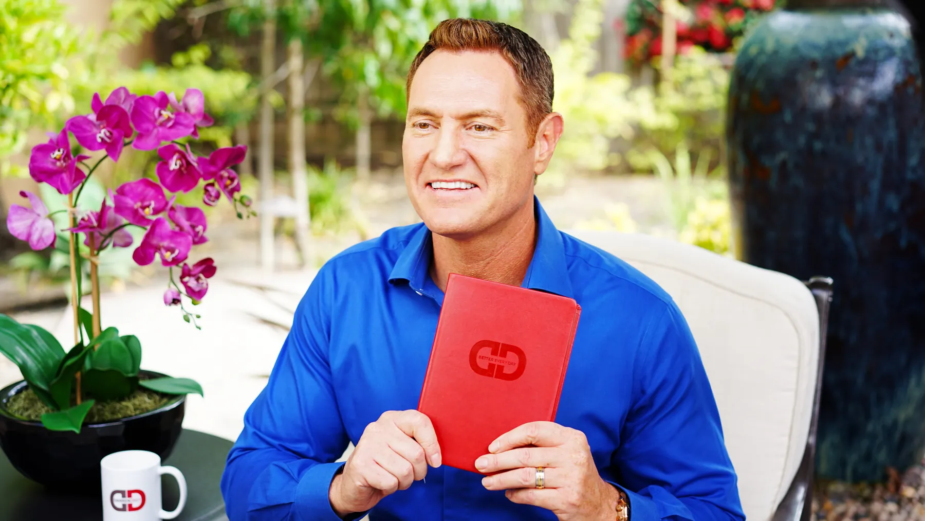 Darren Hardy seated on the DarrenDaily garden set holding the red DarrenDaily journal