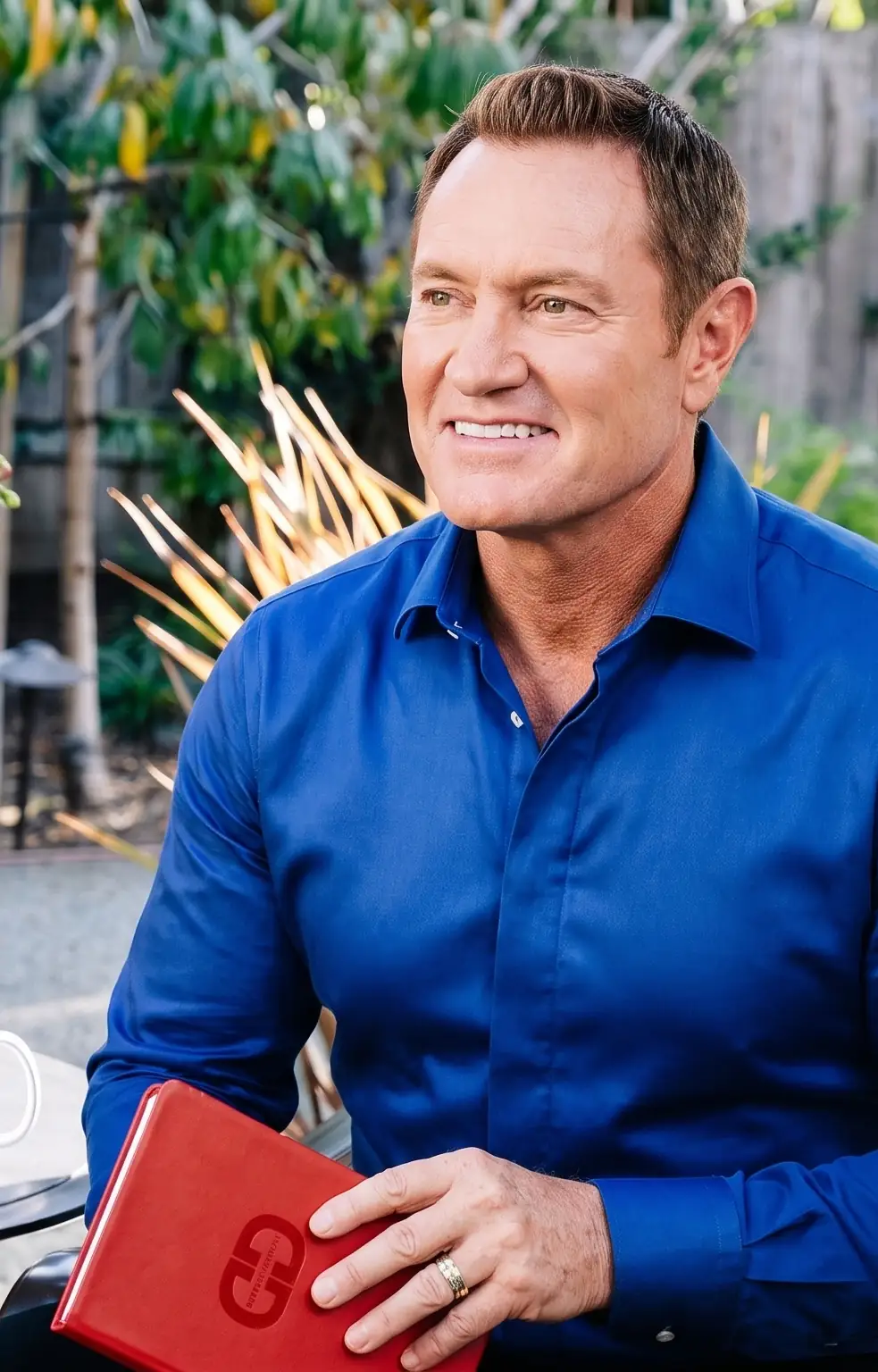 Darren Hardy seated on the DarrenDaily garden set holding the red DarrenDaily journal
