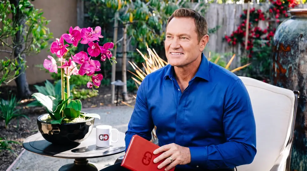 Darren Hardy seated on the DarrenDaily garden set holding the red DarrenDaily journal