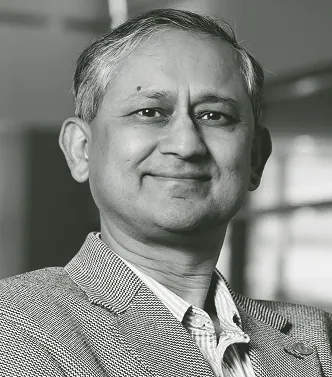 Smiling middle-aged man in a checkered blazer and collared shirt with a blurred indoor background.