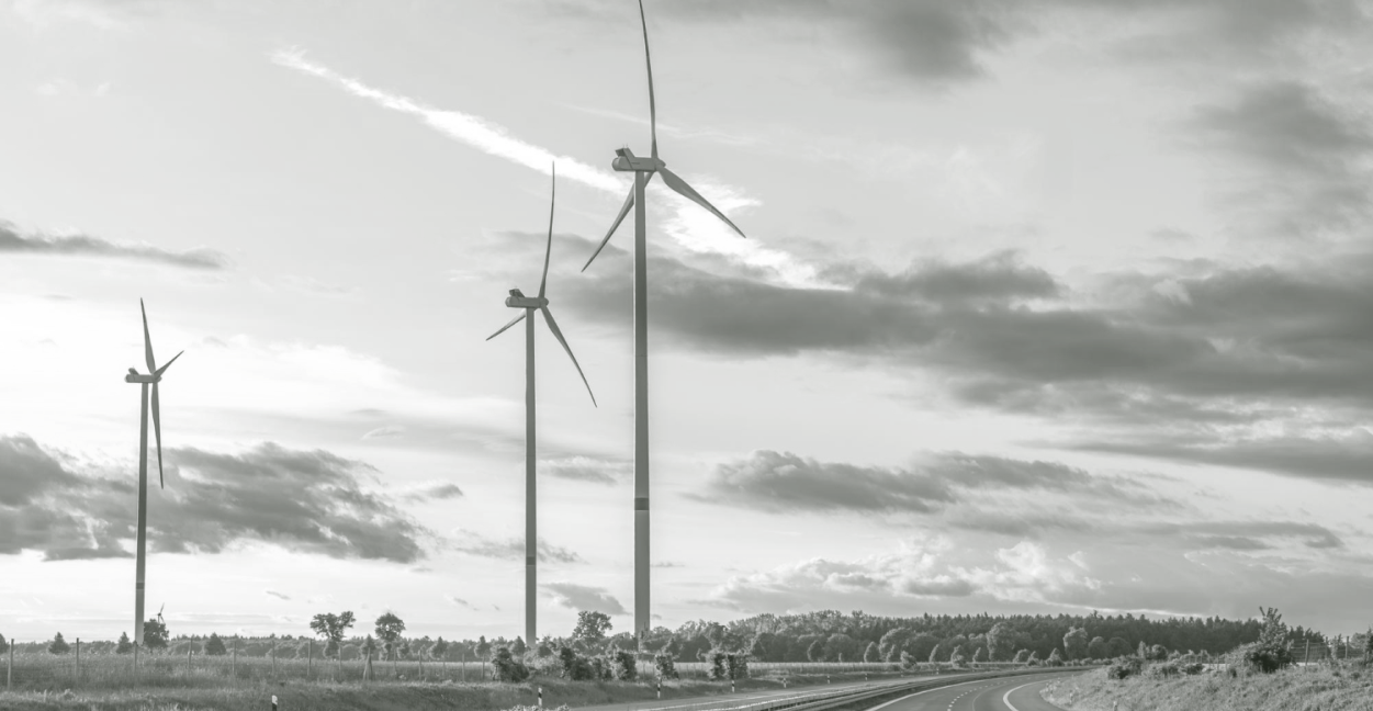 Three wind turbines standing near a curved road under a cloudy sky in a rural landscape.
