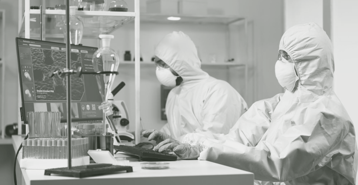 Two scientists in full protective suits working at a lab computer with molecular data on screen.