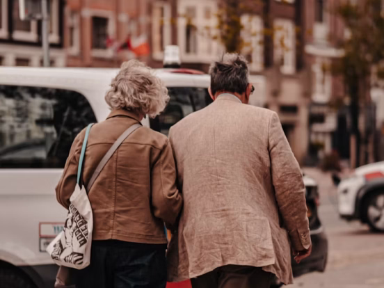 Smiling elderly couple embracing outdoors amid green leafy plants and trees.