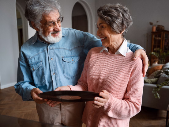 Smiling elderly couple embracing outdoors amid green leafy plants and trees.