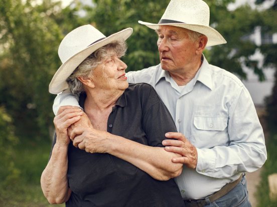 Smiling elderly couple embracing outdoors amid green leafy plants and trees.