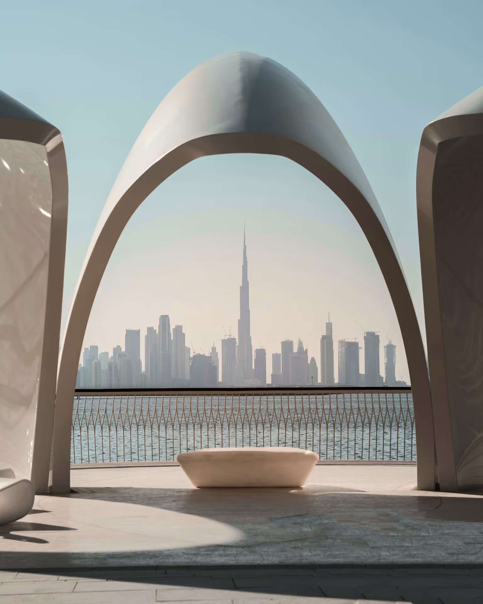 View of Dubai skyline with Burj Khalifa framed by a modern white arch and waterfront railing.