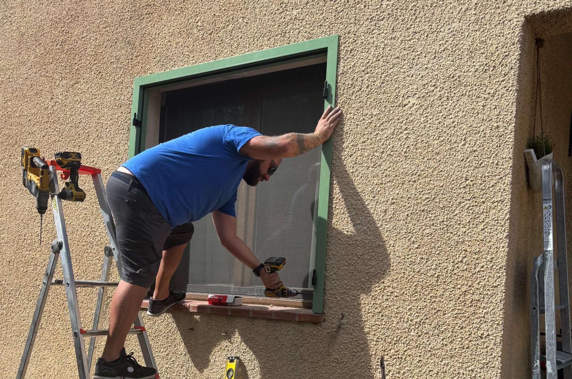 Technicien PRO'PVC posant une moustiquaire sur une fenêtre à volets verts en plein soleil
