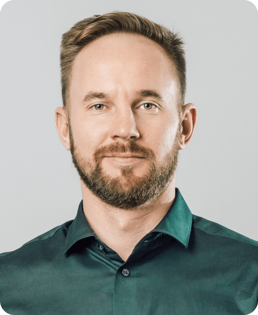 Headshot of a bearded man with short hair wearing a dark green collared shirt against a plain light gray background.