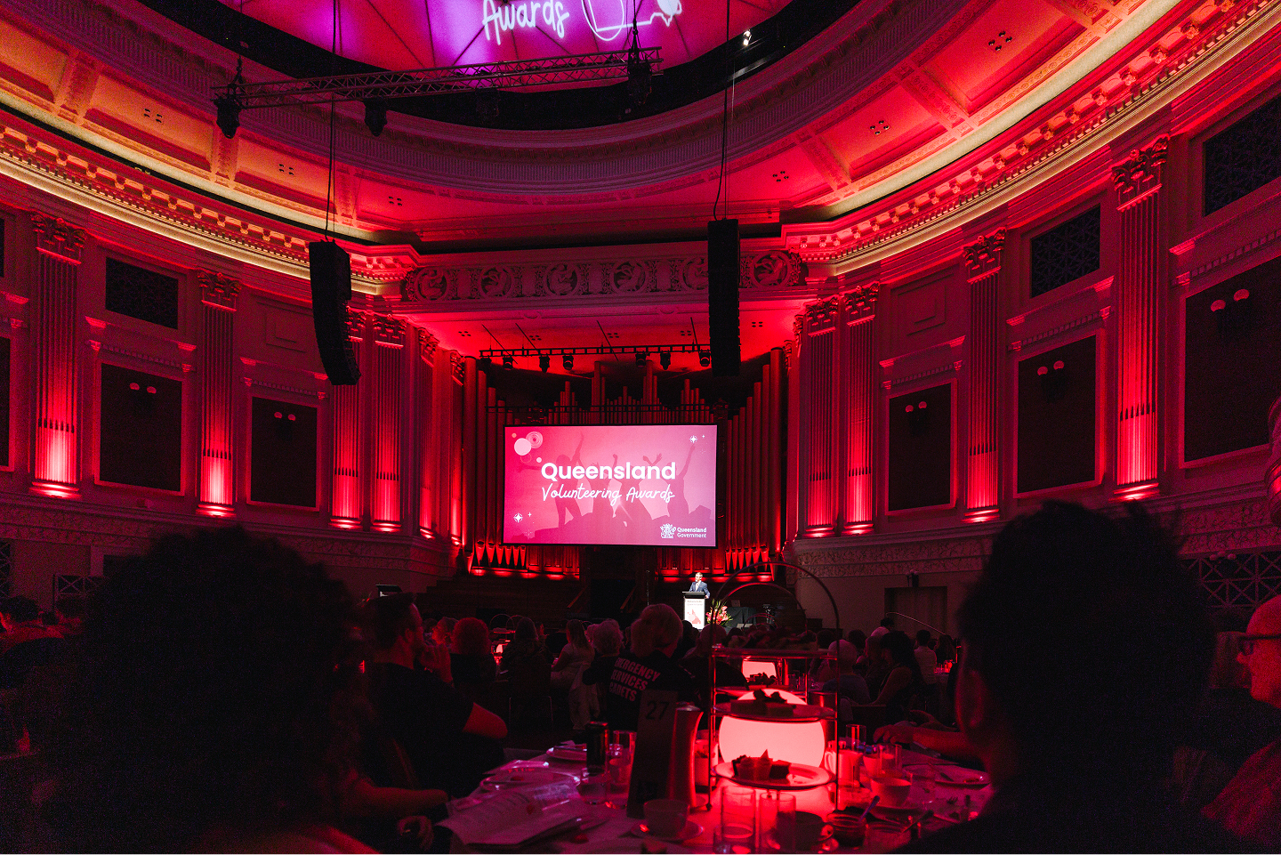 Audience seated in a grand hall with red lighting watching a speaker at a podium during the Queensland Volunteering Awards event.
