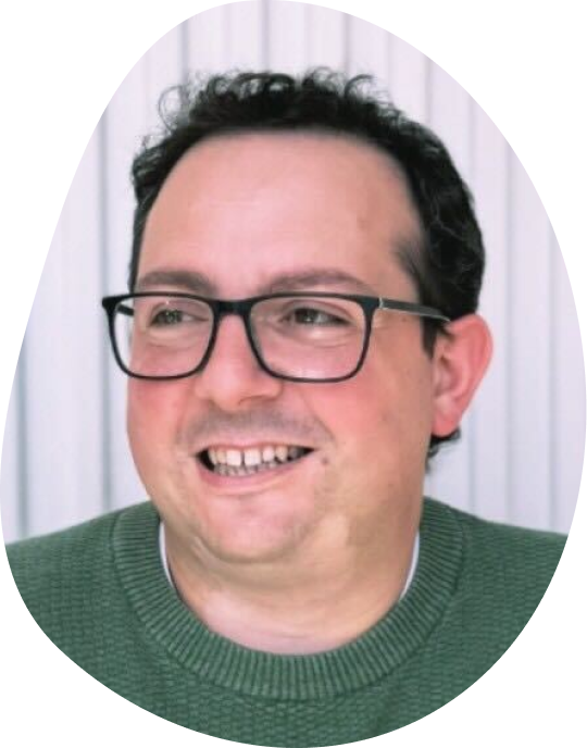 Man with glasses smiling in a green sweater against a white-striped background, exuding a friendly and cheerful mood.