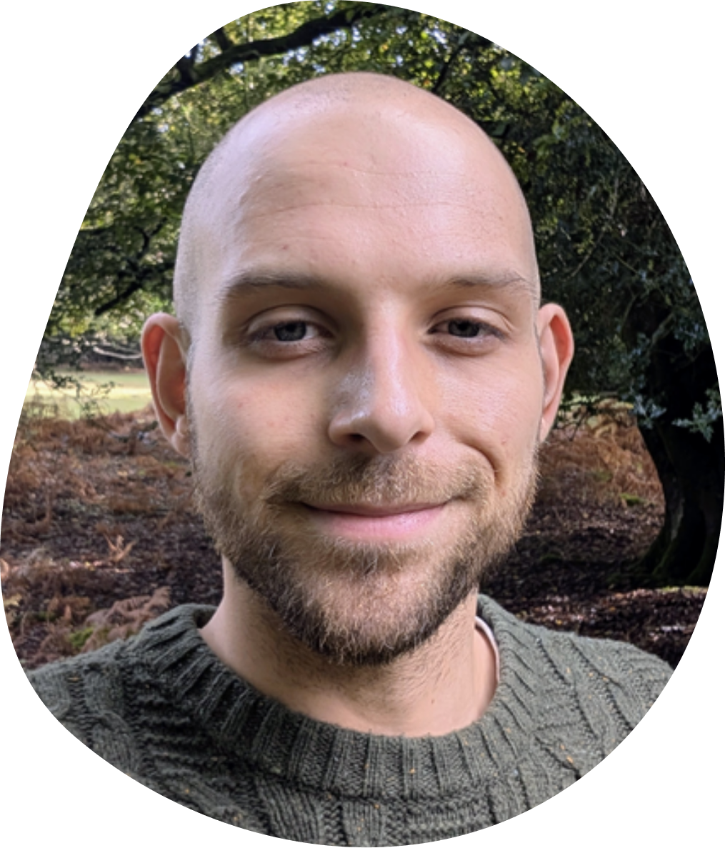 Man with a bald head and beard smiling slightly, wearing a green knit sweater. Background shows a forest with trees and foliage.