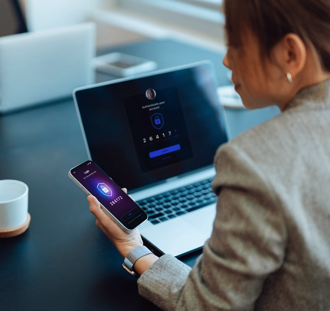 Woman holds phone showing login code while laptop displays same two-factor authentication screen.