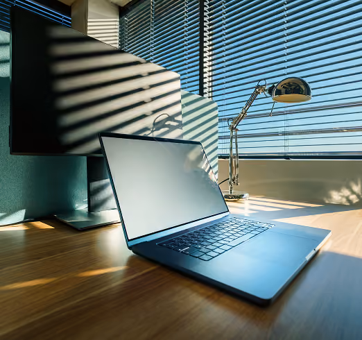 Laptop and monitor on a wooden desk lit by sunlight coming through window blinds with a desk lamp nearby.