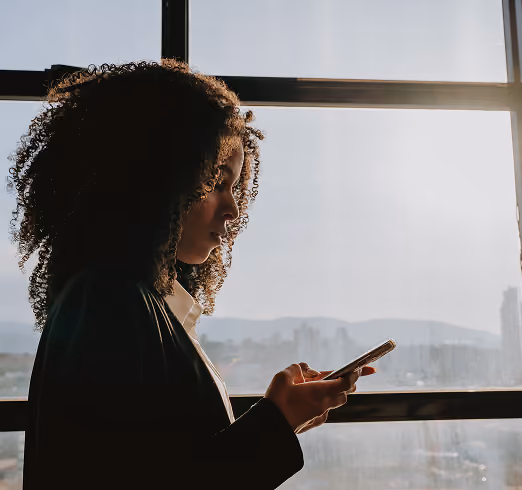 Profile of woman with curly hair looking at her smartphone by a large window in daylight.