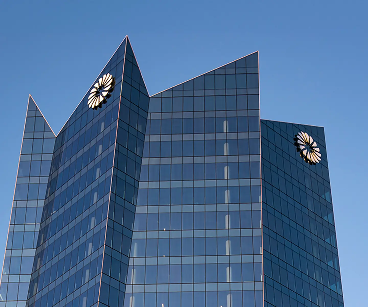 Modern glass office building with two flower-shaped logos near the top against a clear blue sky.
