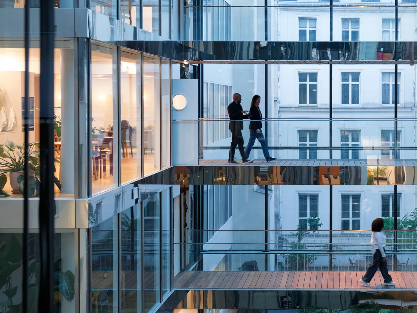 Two people walking inside a modern glass office building on upper levels and one walking below on a wooden floor.
