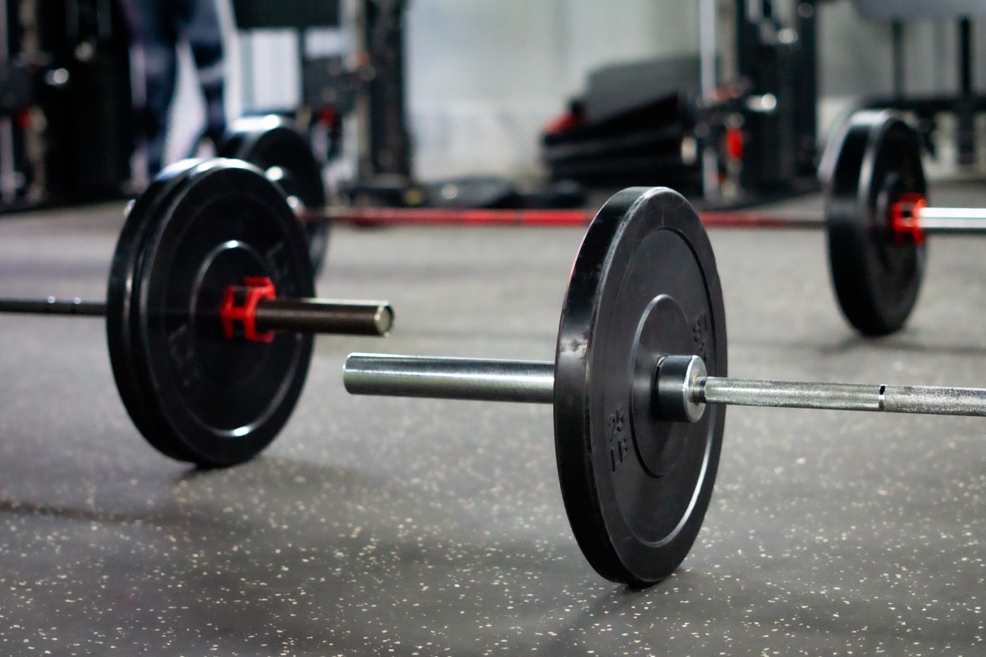 A close-up view of barbells loaded with black bumper plates and red collars resting on a protective speckled rubber floor, ready for use.