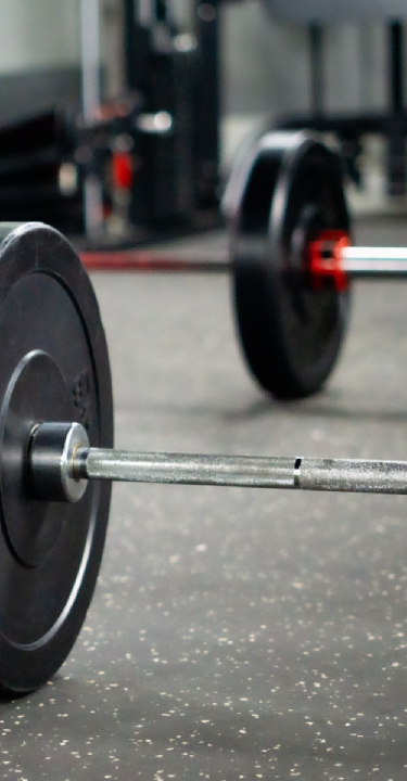 A close-up view of barbells loaded with black bumper plates and red collars resting on a protective speckled rubber floor, ready for use.