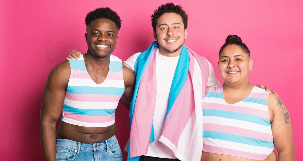 Three smiling individuals wearing trans pride crop tops and holding a trans pride flag against a pink background.