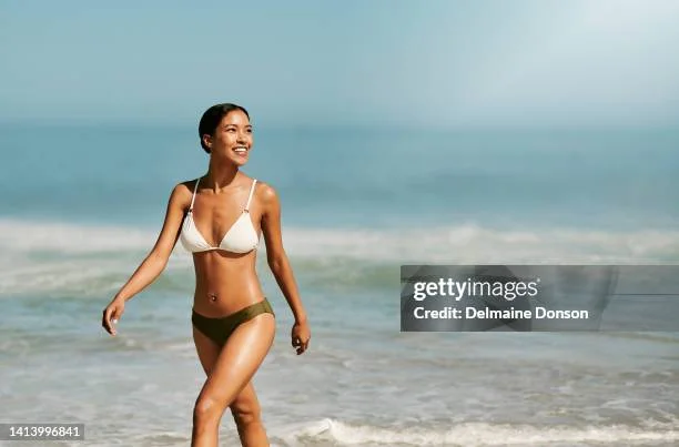 Woman in a white bikini walking on the beach, smiling under the sun.