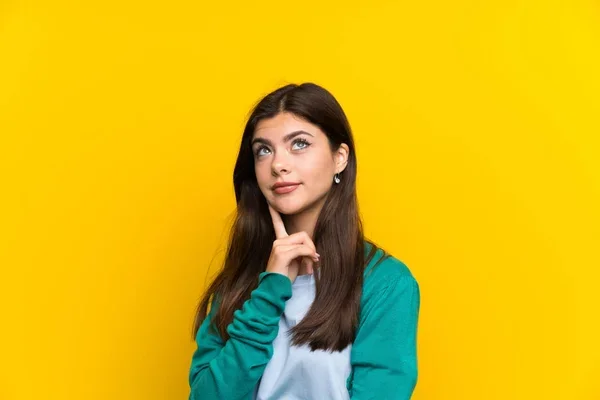 A thoughtful young woman in casual attire standing against a bright yellow background, touching her chin and looking upward.