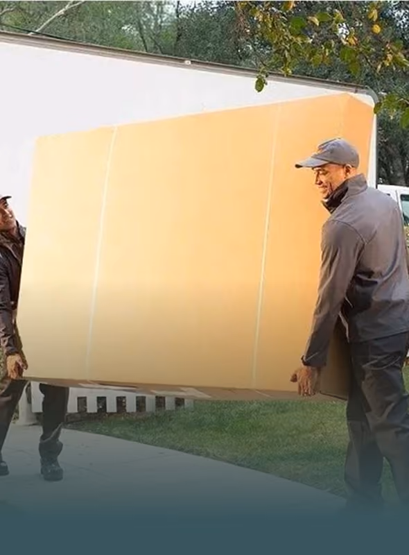 Two men in caps carrying a large cardboard box outdoors near a white truck and trees.