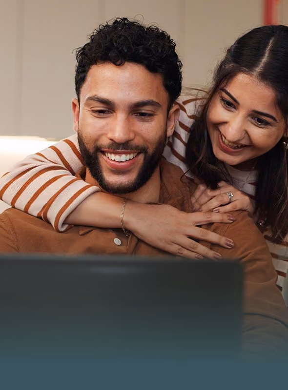 Smiling couple looking at a laptop screen together, woman hugging man from behind.