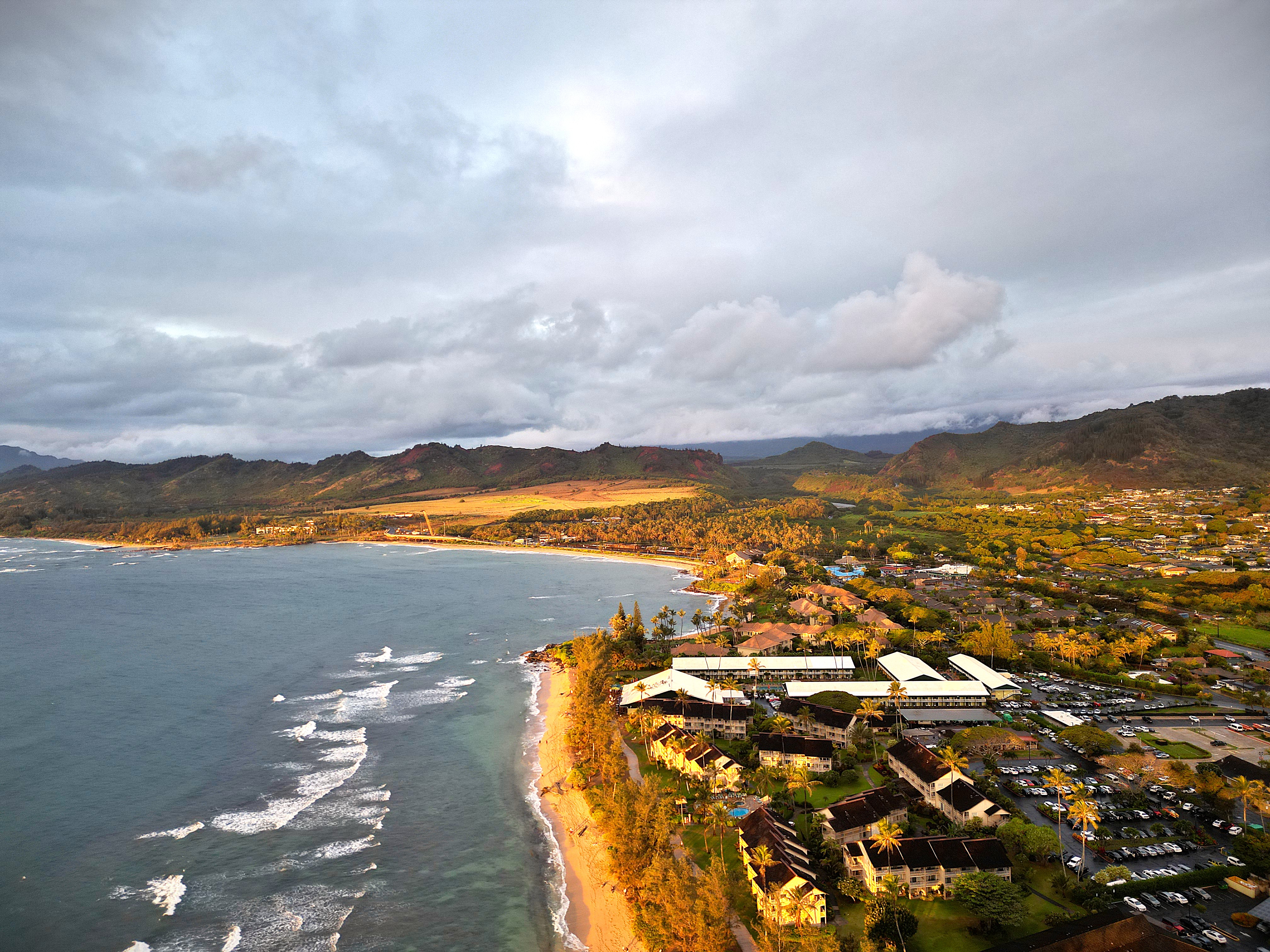 Aerial drone view of a Kauai resort coastline glowing at golden hour, mountains in the background.