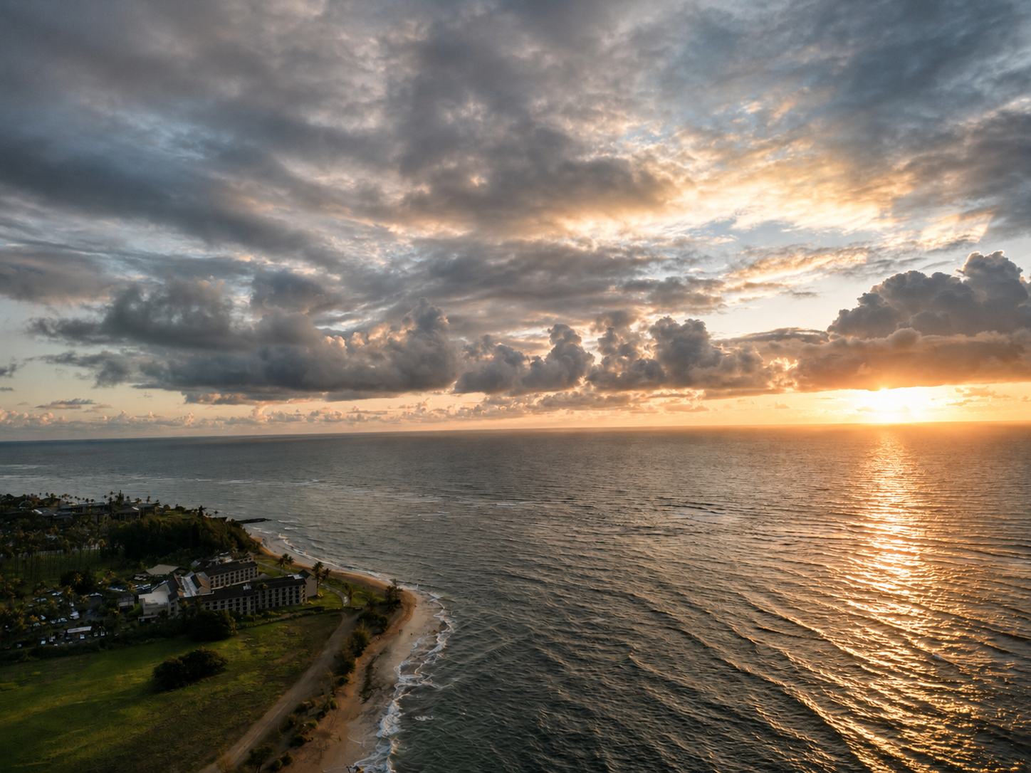 Aerial drone view of a golden sunset over the ocean along a Kauai resort coastline.