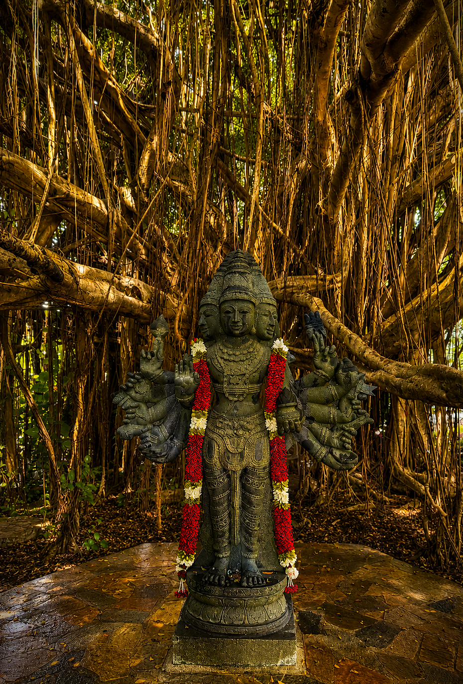 Multi-armed deity statue draped in flower leis beneath a banyan tree on Kauai.
