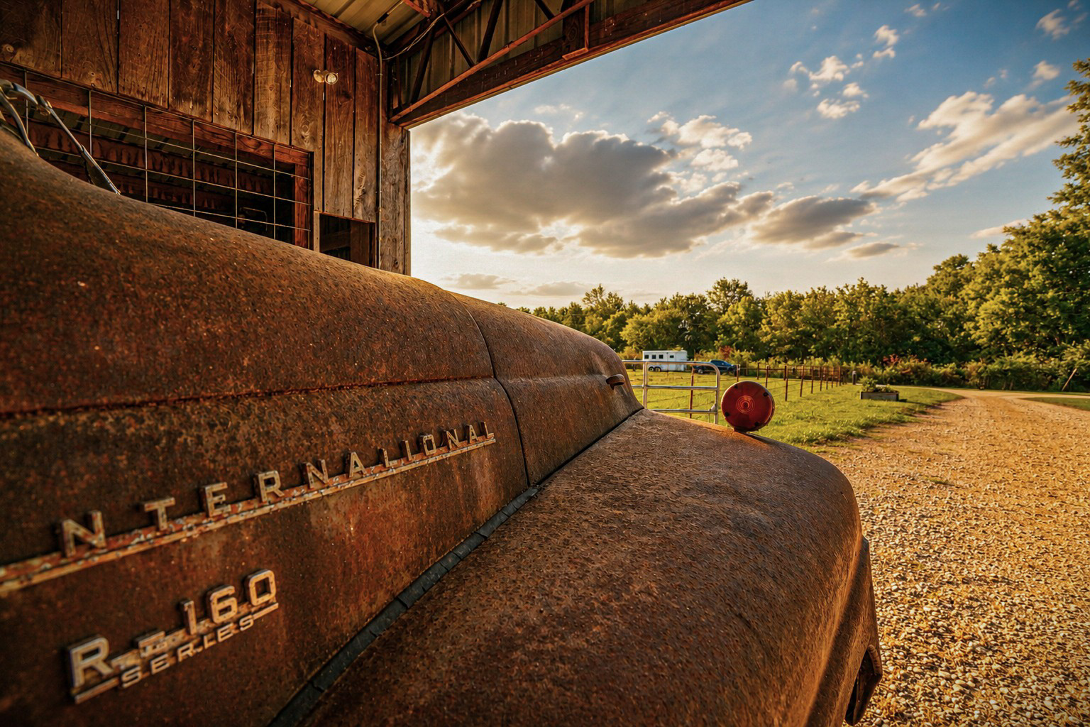 Close-up of a rusted vintage International R-160 truck's hood and fender inside the barn, overlooking the gravel drive at sunset.