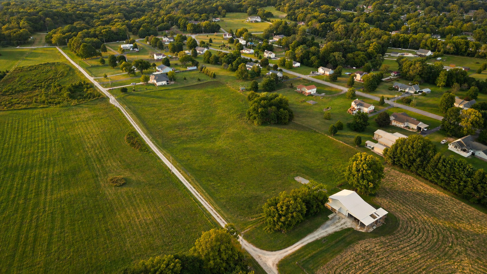 Aerial view of a paved road leading from open pastureland into a Middle Tennessee neighborhood, with a white pole barn visible on the property at right.