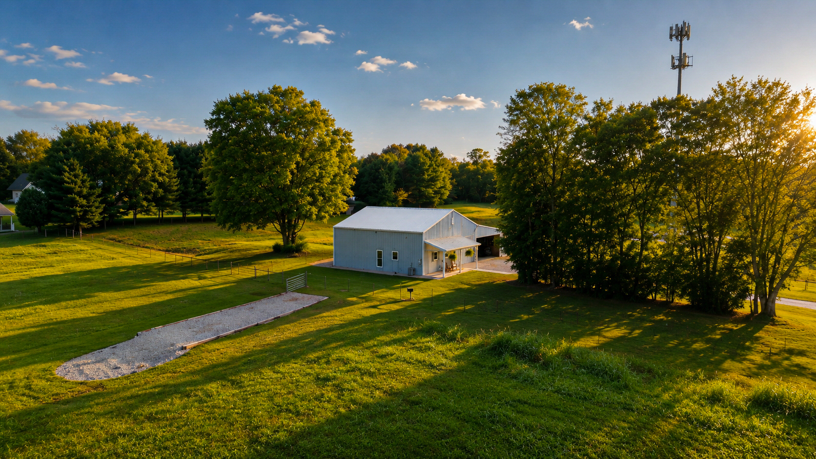 Elevated aerial view of the white pole barn and pasture at golden hour, with a cell tower and tree line in the background.