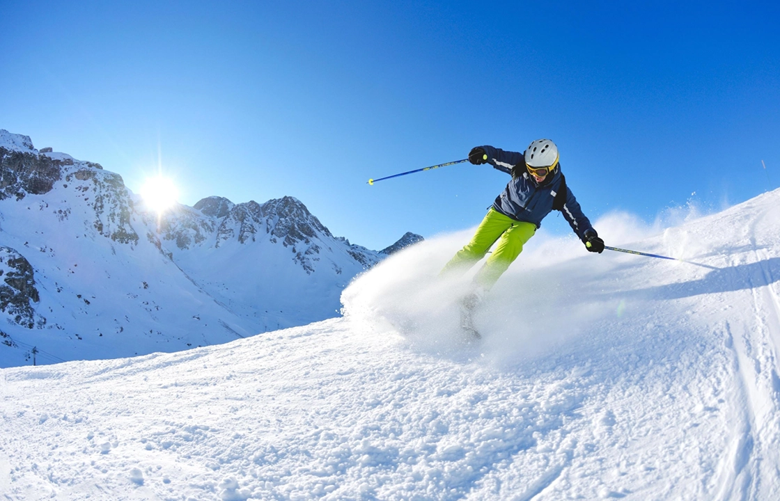 Skier in green pants and helmet carving snow on a sunny mountain slope.