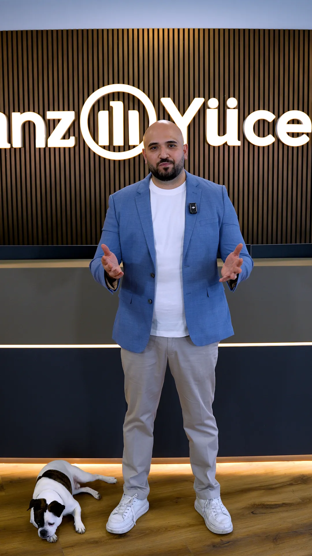 Versicherungsberater Özgün Yücel aus Köln in blauem Sakko und weißem T-Shirt steht vor einer Wand mit Allianz-Lichtschrift, Hund liegt am Boden.