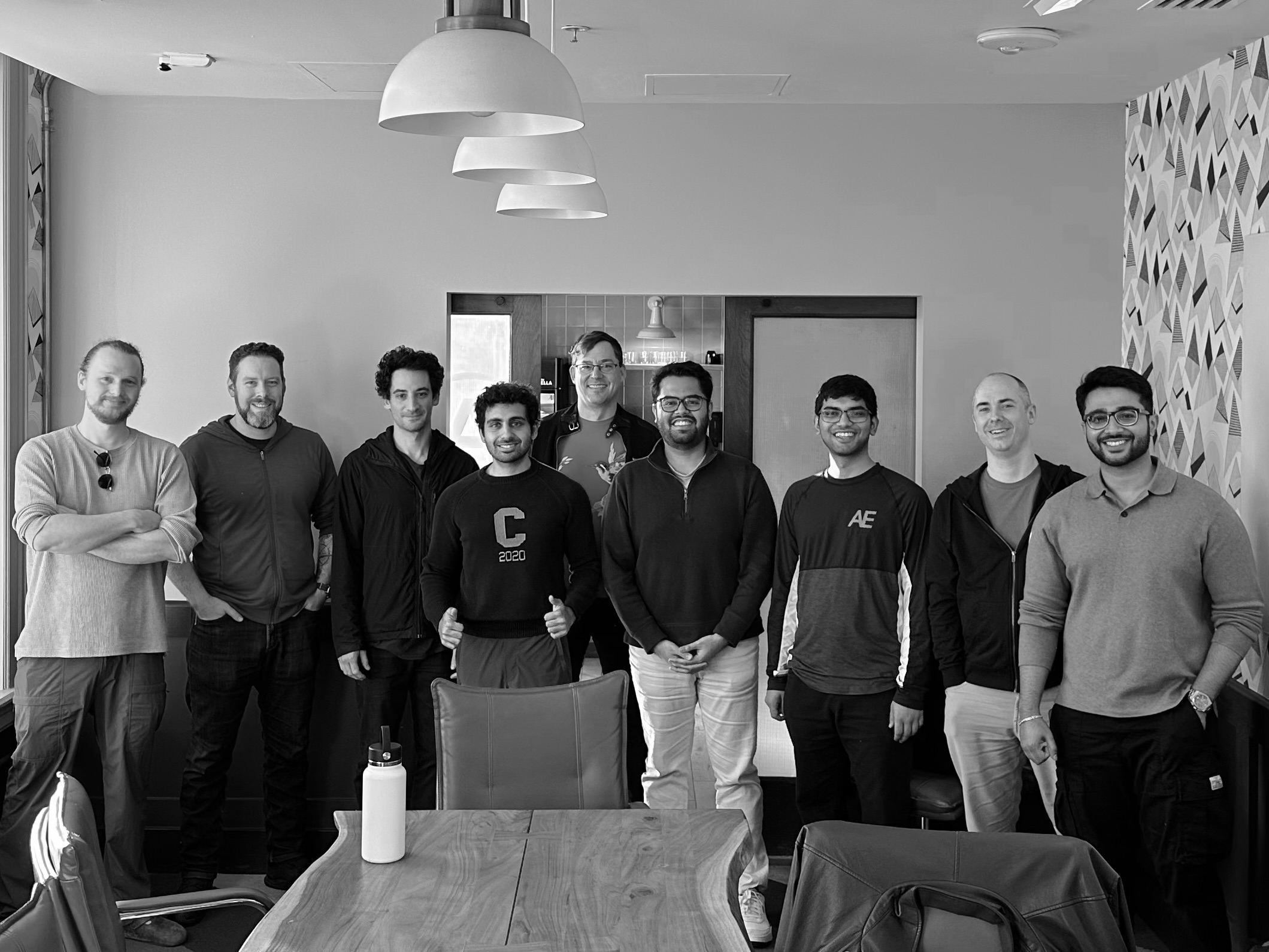 Group of nine men standing together in a room behind a wooden table with chairs and hanging lights.