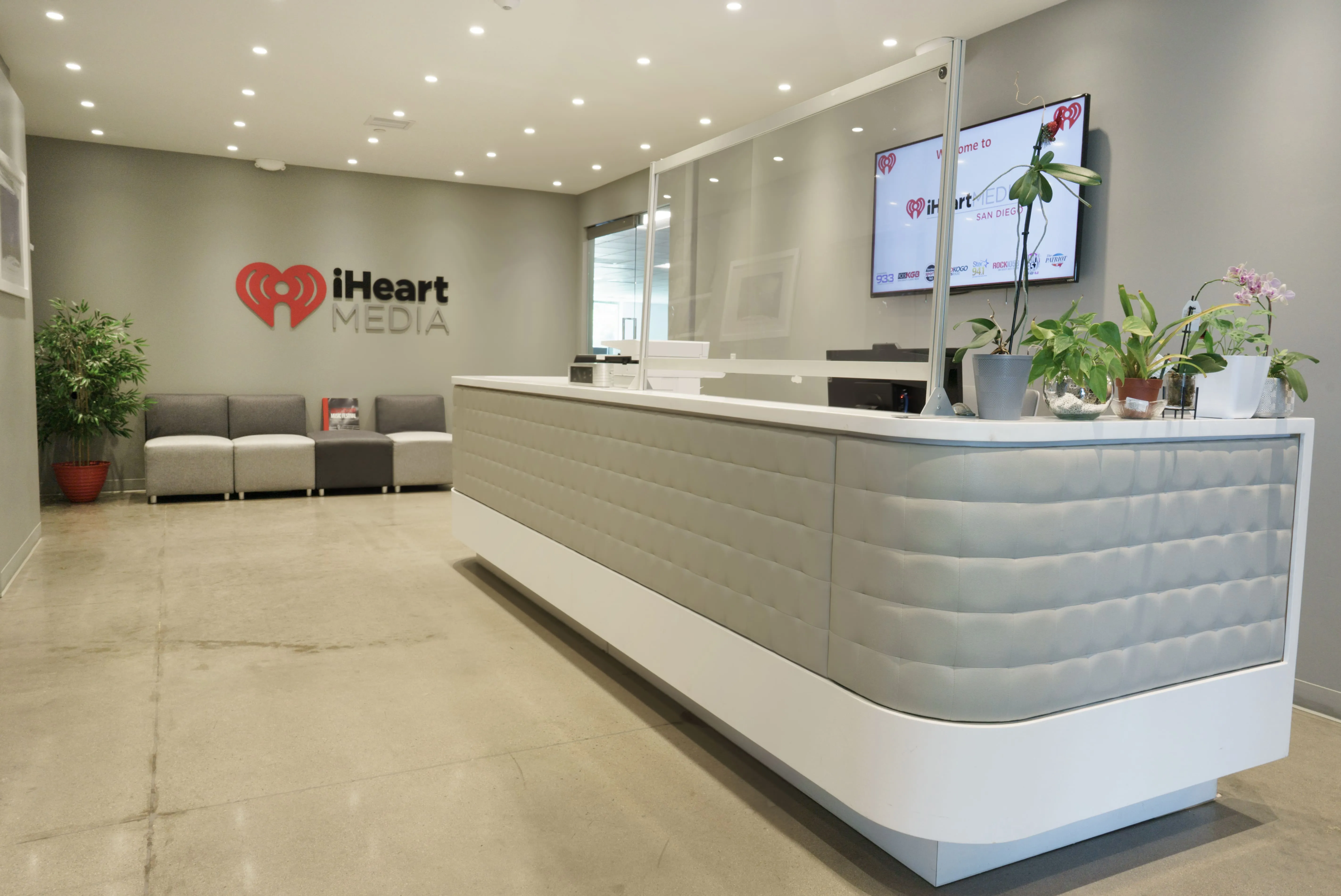 Reception desk with plants and clear divider in iHeart Media office lobby with company logo on wall.