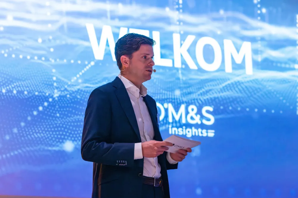 Man in pak met microfoon spreekt op een evenement met een blauw scherm met 'WELKOM' op de achtergrond.
