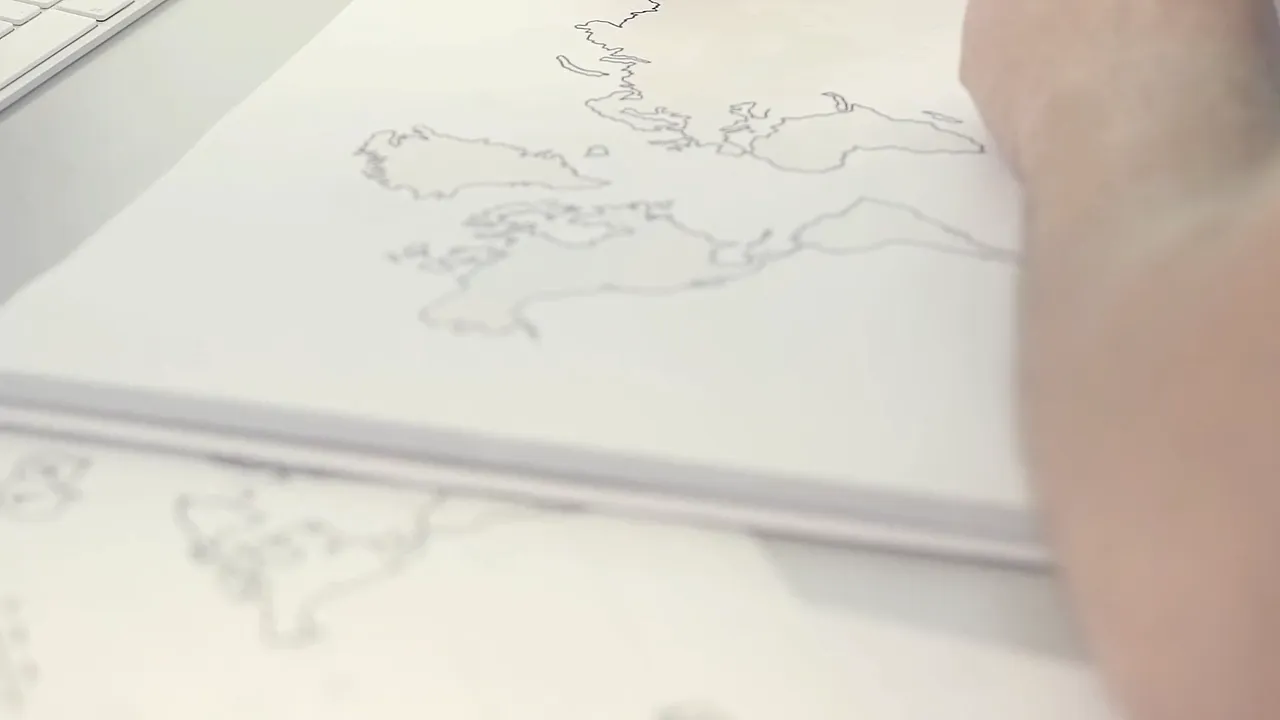 Close-up van een hand die een wereldkaart schetst op papier naast een toetsenbord.