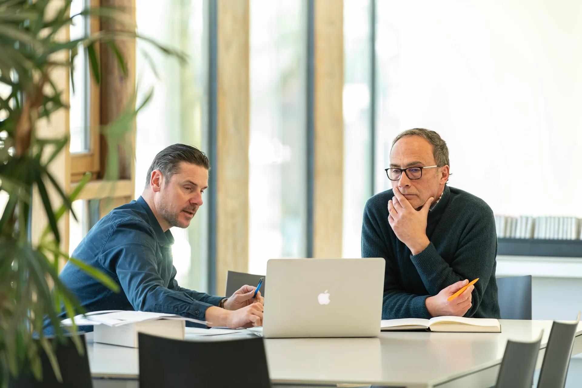 Twee mannen zitten aan tafel, kijken geconcentreerd naar een laptop met notitieboekjes en pennen.