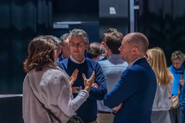 Groep volwassenen praat tijdens een evenement in een binnenruimte met donkere muren, waarbij drie personen in het midden in gesprek zijn en anderen op de achtergrond staan.