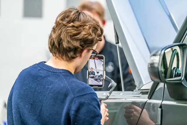 Een man met een blauwe trui filmt een andere man met zijn smartphone op een gimbal, terwijl deze laatste voor een open motorkap van een voertuig staat.