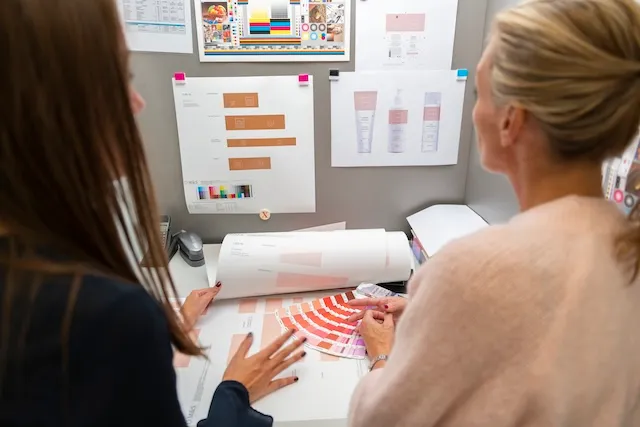 Twee vrouwen bekijken kleurstalen en grafische ontwerpen op een bureau met afdrukken aan een grijze wand.