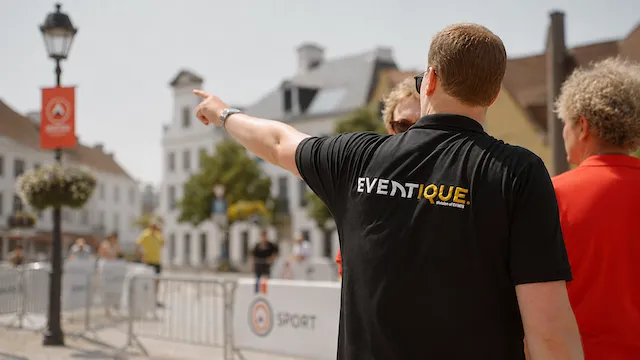Een man met een zwarte Eventique polo wijst naar links in een buitenomgeving met gebouwen, een lantaarnpaal en enkele personen op de achtergrond.