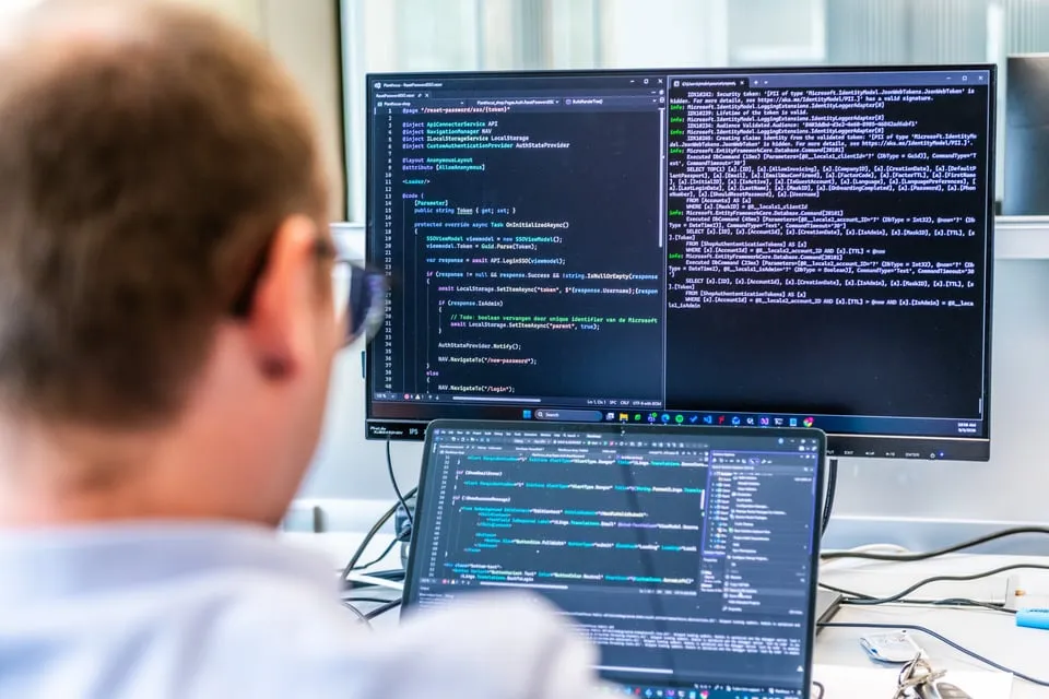 Persoon met bril werkt op een computer met twee schermen waarop code en foutmeldingen worden weergegeven.