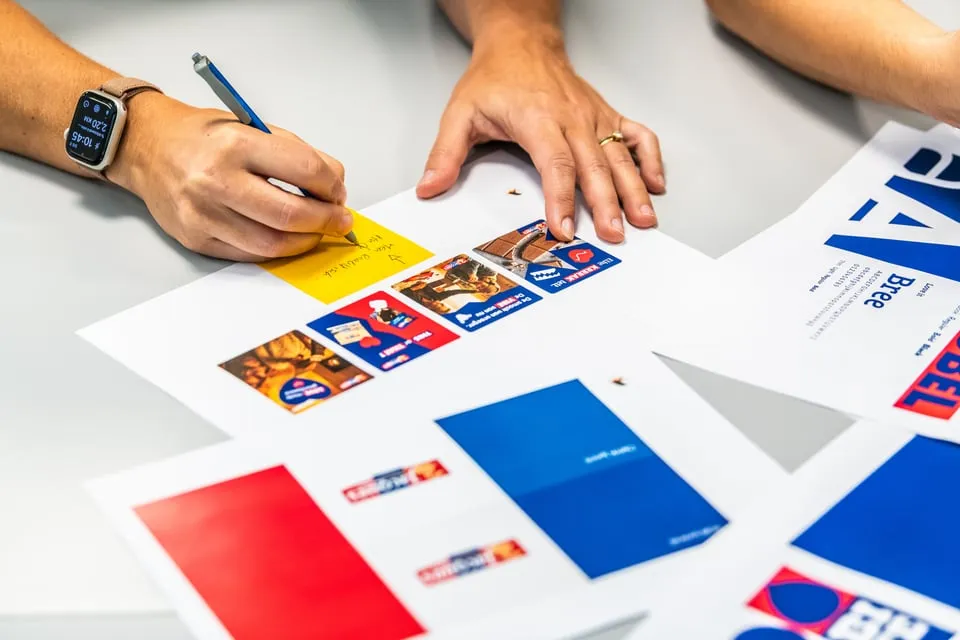 Twee handen van een persoon aan een bureau met verschillende pagina's waarop afbeeldingen en logo's in rood en blauw zijn gedrukt; de linkerhand schrijft iets op een gele notitie met een blauwe pen, de rechterhand ligt op de tafel met een gouden ring aan de ringvinger.
