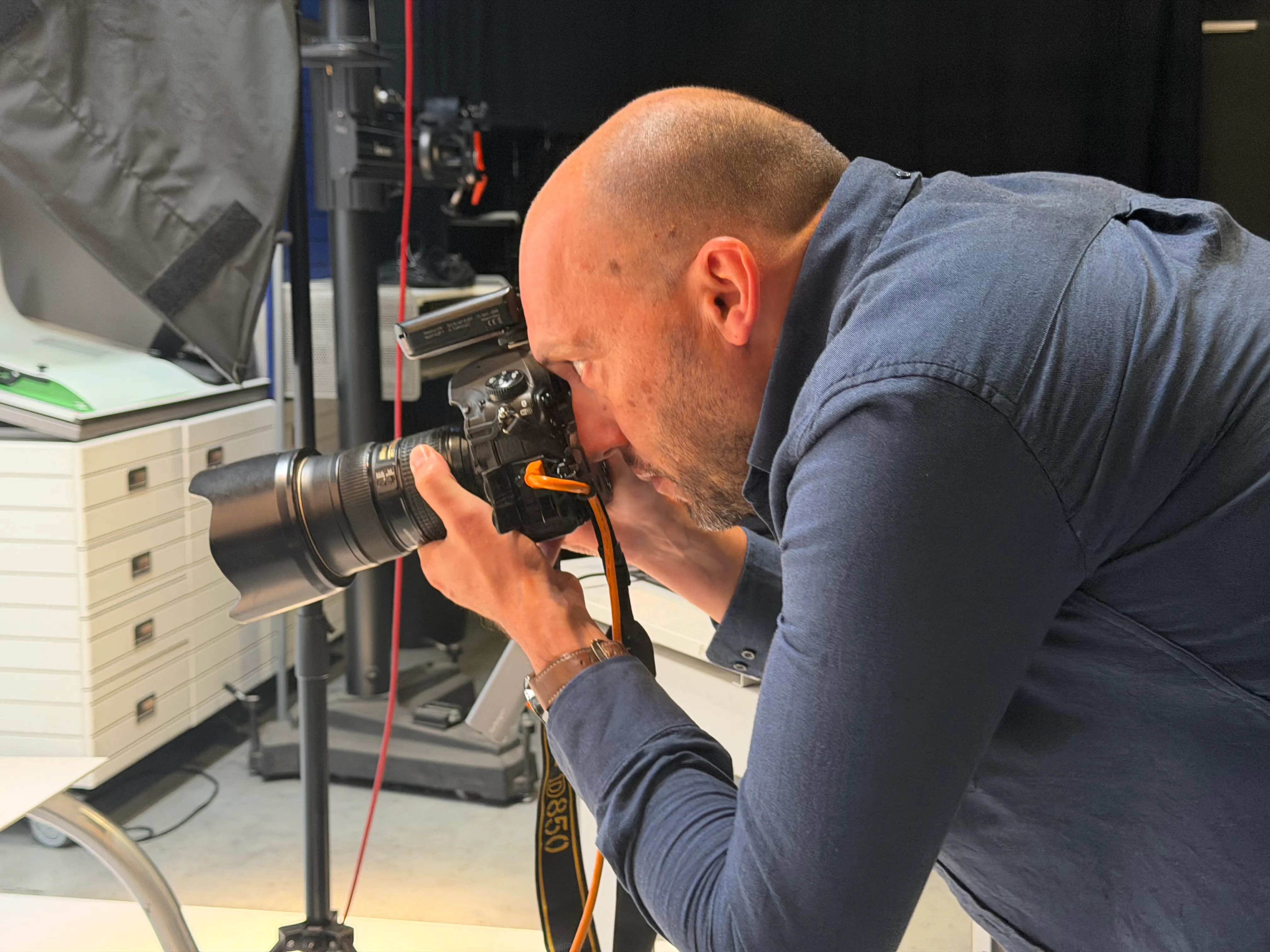 Een man met een blauwe lange mouwen shirt buigt voorover en richt een professionele camera met een grote lens op een onzichtbaar onderwerp in een studioruimte.