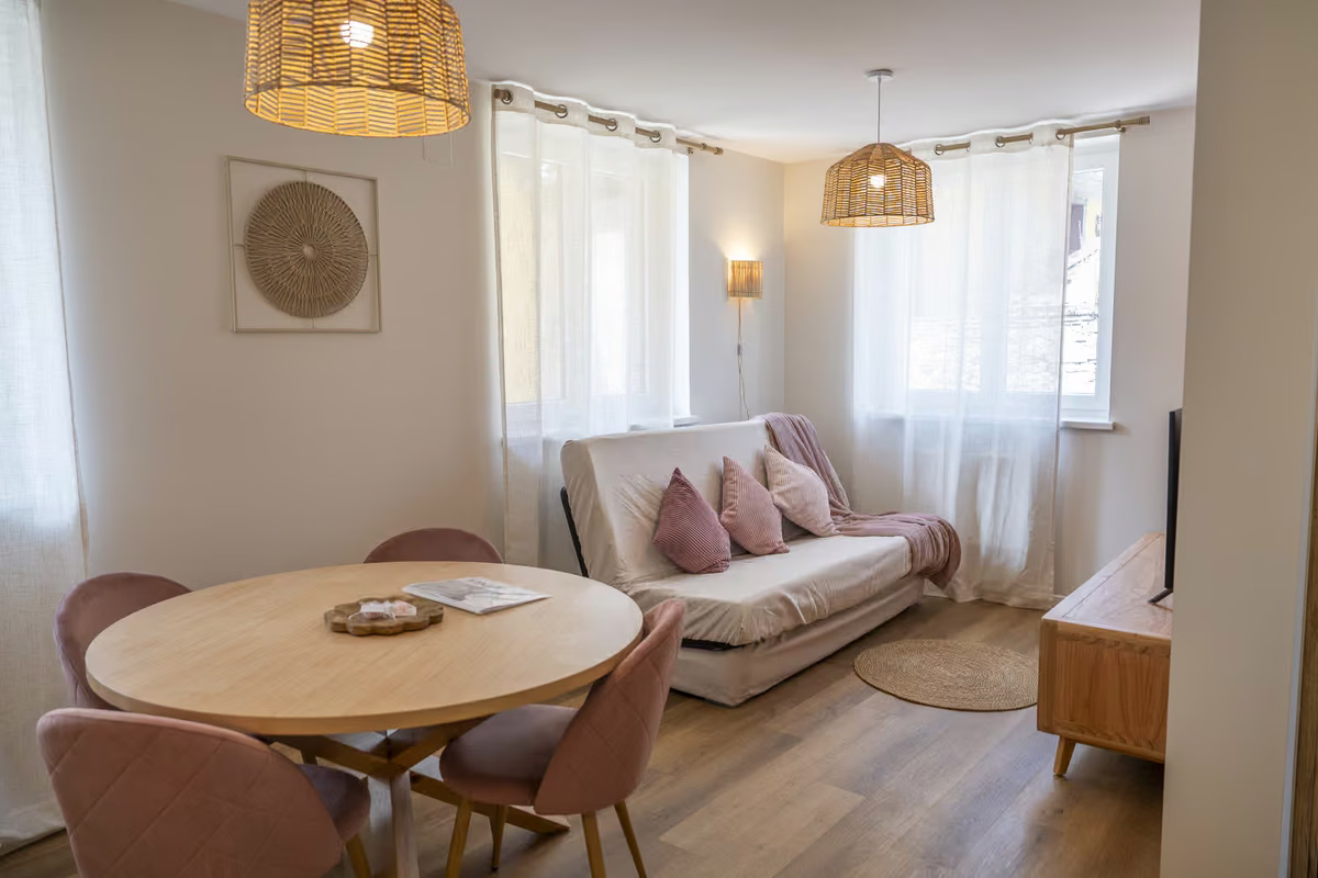 Pièce de vie lumineuse d'un gîte avec table ronde, chaises roses et canapé convertible.
